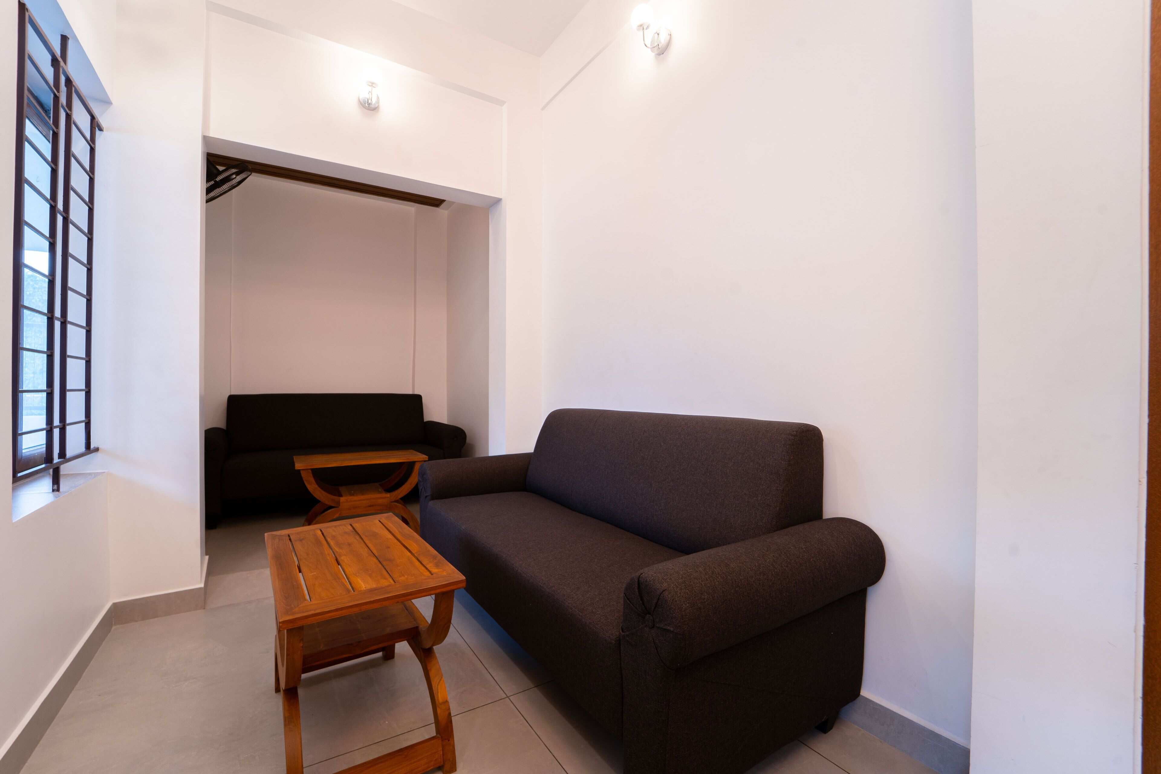 A modern living room with a dark brown sofa, a wooden coffee table, and a small table with chairs. There is a window with a metal grid on the left and a door leading to a closet.
