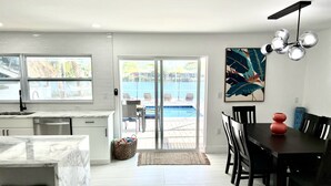 Une cuisine moderne avec un plan de travail en marbre, un coin repas avec une table et des chaises en bois foncé, et une vue sur une piscine à travers des portes vitrées.