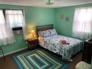 Quarto com cama, mesa de cabeceira com abajur, radiador e janela com cortinas.