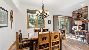 Een eethoek met een houten tafel en stoelen, een kroonluchter, een open haard en grote ramen.