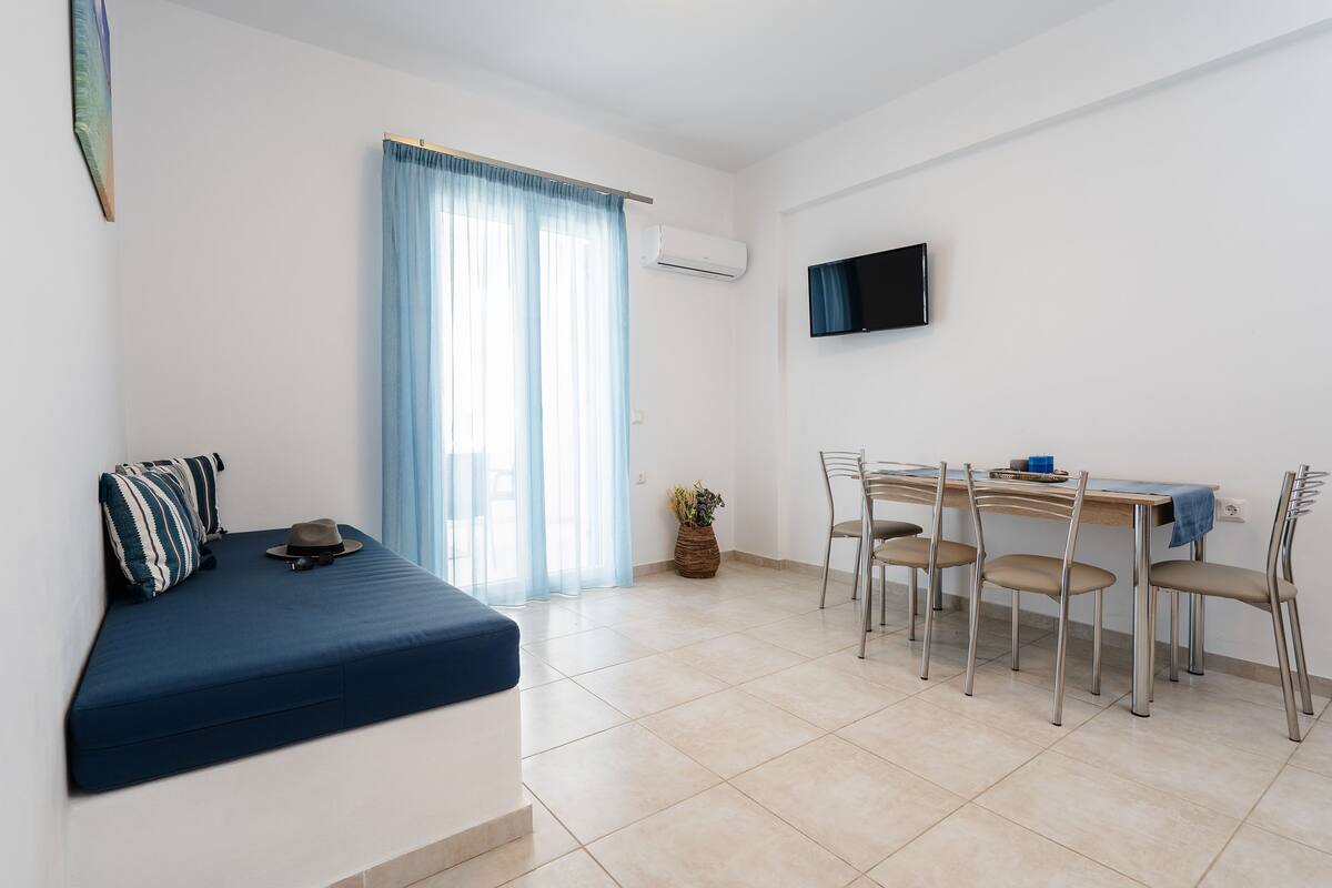 A room with a bed, a dining table with chairs, a TV, and a window with curtains.