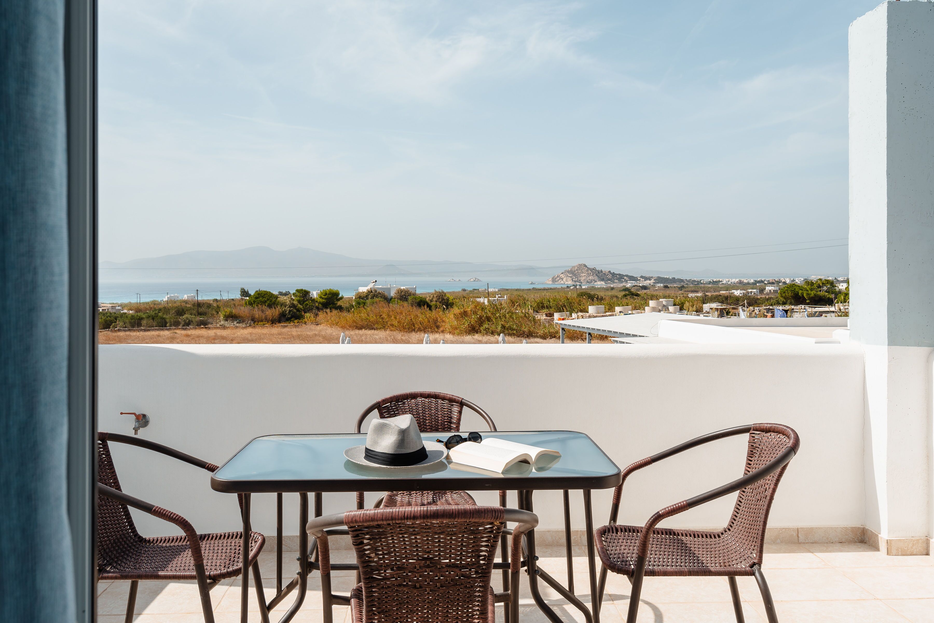 A balcony with wicker chairs, a table, and a view of the sea and distant landscape.