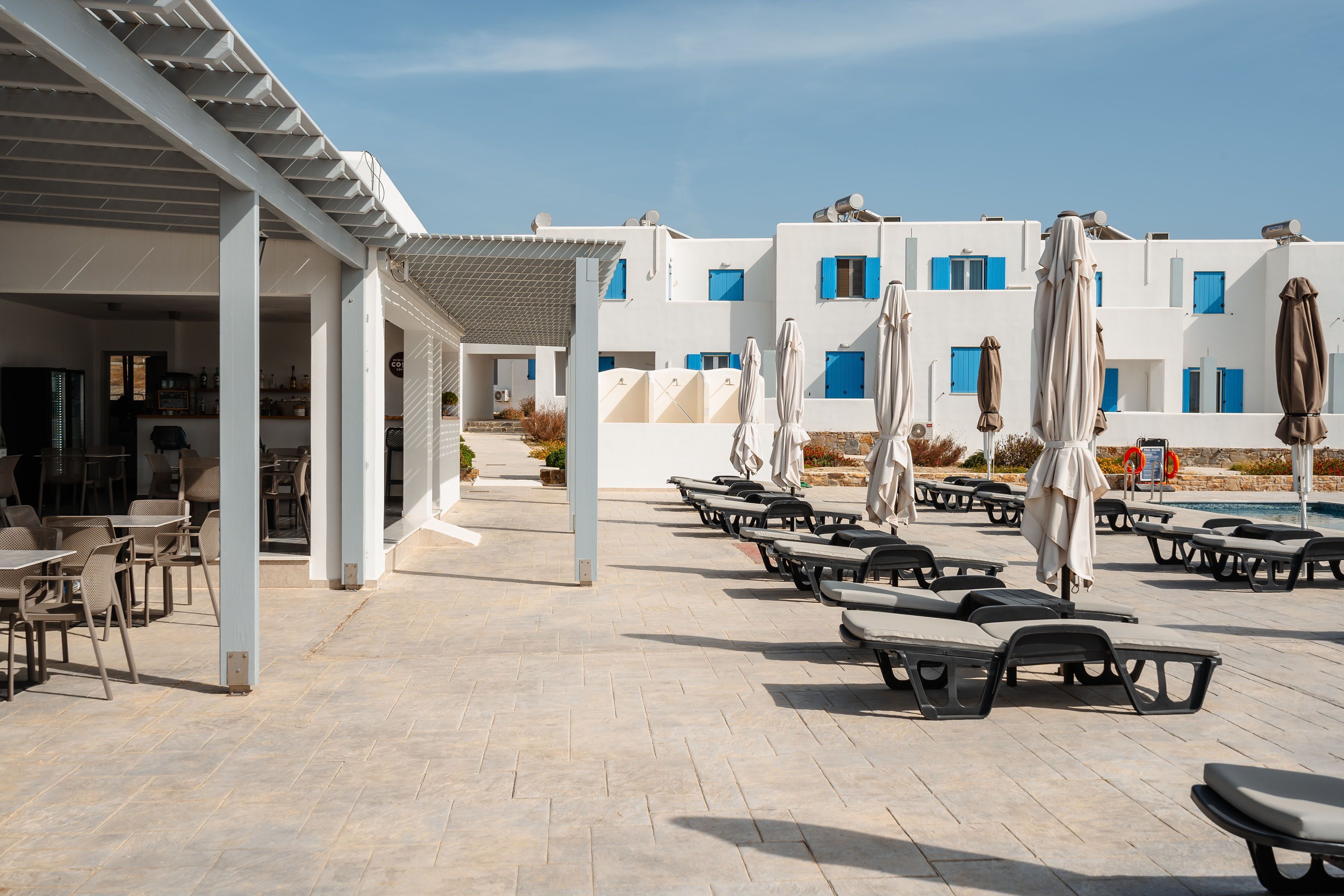 A covered outdoor seating area with tables and chairs, umbrellas, and a pool in the background.