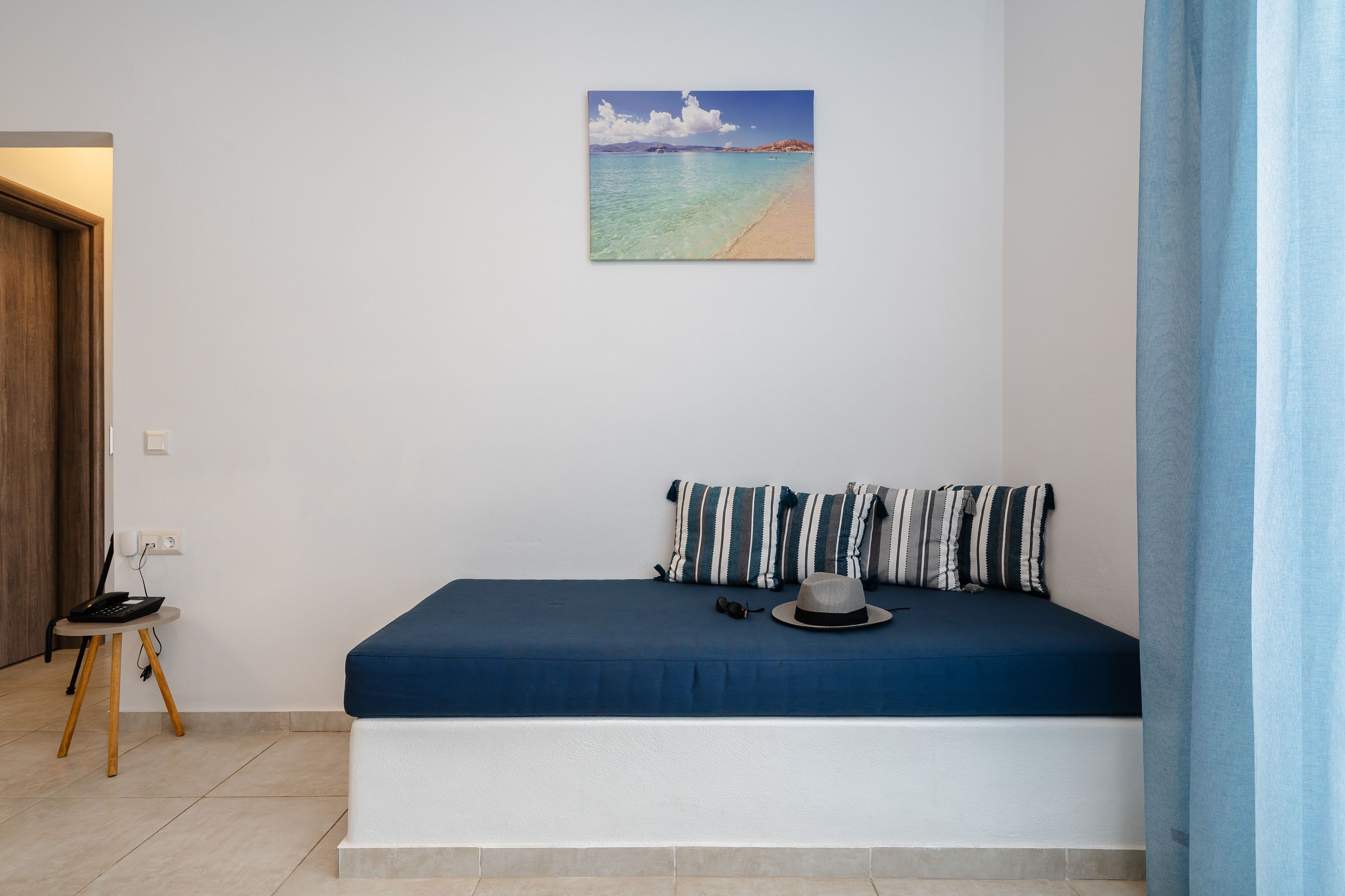A bedroom with a bed, a small table, a chair, a hat, and a painting of a beach.
