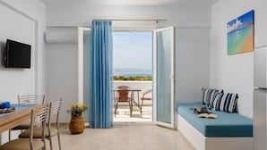 Una habitación con una mesa de comedor, sillas, un sofá con cojines, un televisor y vista al mar a través de una puerta corredera.