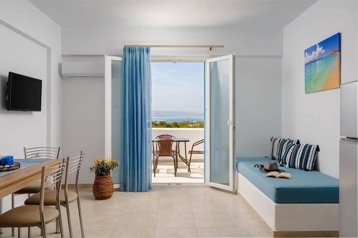 A room with a dining table, chairs, a sofa with cushions, a TV, and a view of the sea through a sliding door.