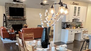 A dining area with a wooden table set for four, surrounded by chairs. A black vase with white flowers is on the table. - The Lighthouse- A Beautiful and Luxurious SmartHome. (Terre Haute)