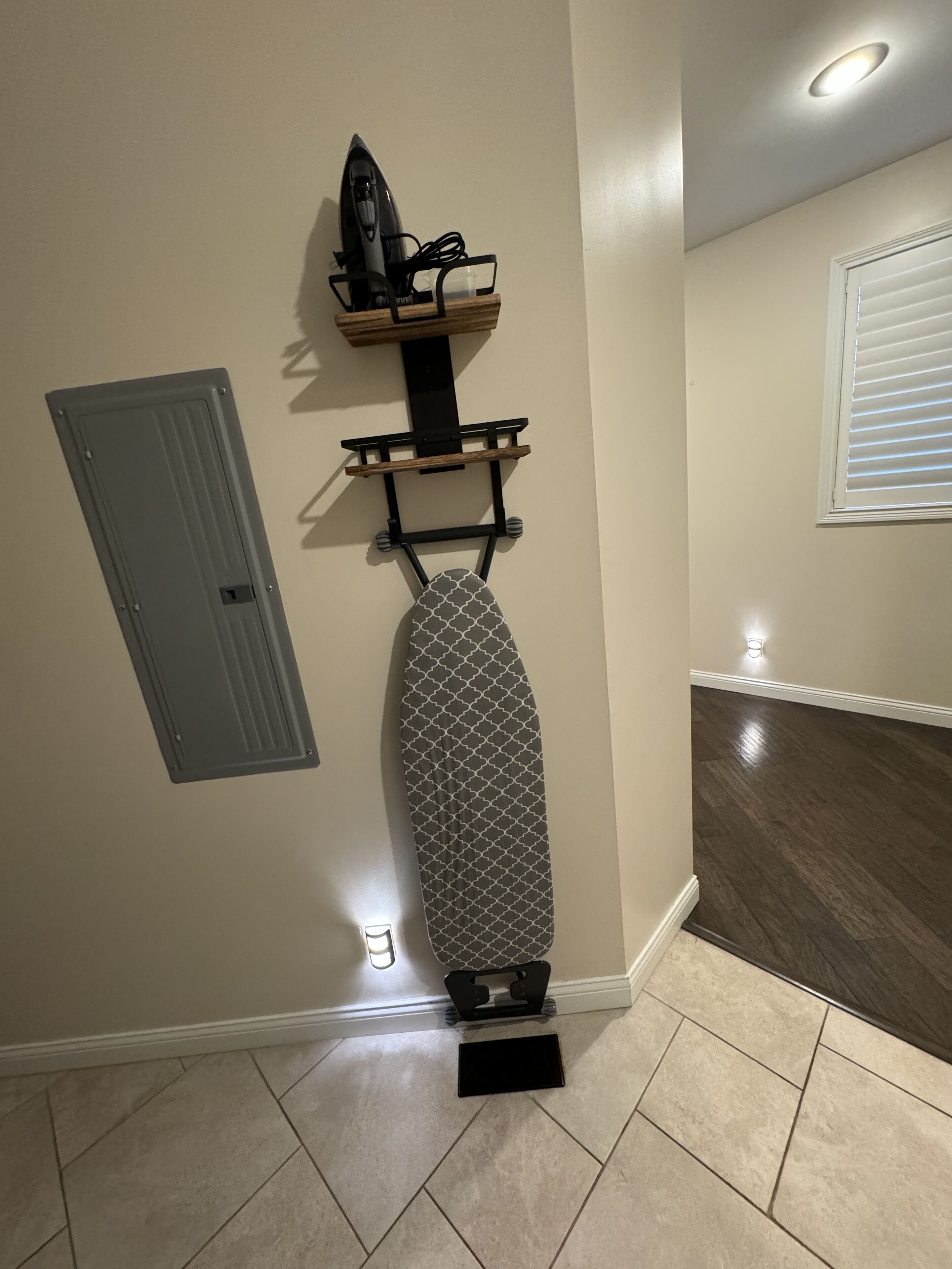 A corner of a room with a wooden floor, a grey door, and a wall-mounted ironing board with an iron and a small shelf above it.