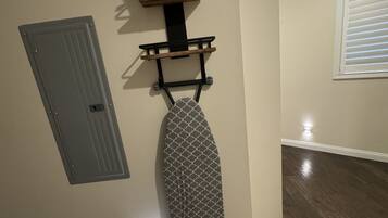 A corner of a room with a wooden floor, a grey door, and a wall-mounted ironing board with an iron and a small shelf above it.