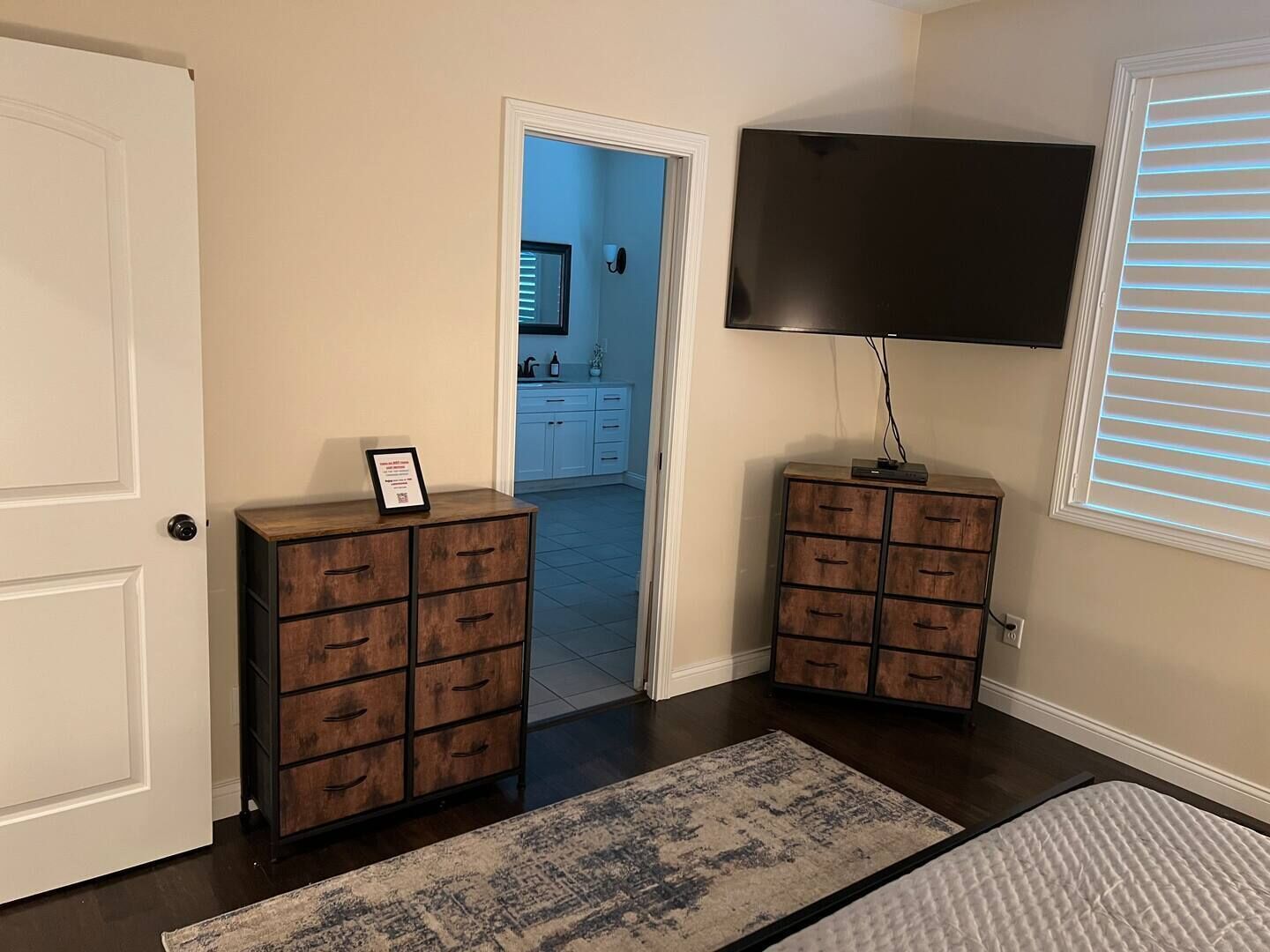 A room with a large flat-screen TV mounted on the wall, wooden cabinets with drawers, a patterned rug, and a door leading to a kitchen area.