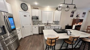 A modern kitchen with white cabinets, a stainless steel refrigerator, a built-in oven, and a dishwasher. There is a dining area with a white island and bar stools. - The Lighthouse- A Beautiful and Luxurious SmartHome. (Terre Haute)