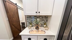 A kitchen with white cabinets, a wooden door, and a countertop with a decorative arrangement including a vase, bottles, and a wooden board. - The Lighthouse- A Beautiful and Luxurious SmartHome. (Terre Haute)