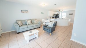 A living room with a sofa, armchair, and coffee table, a kitchen in the background, and a dining area with chairs.