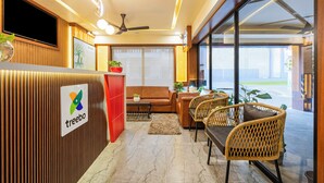 A modern reception area with a wooden reception desk, a brown sofa, wicker chairs, and a large window offering a view of the outdoors. - Treebo Premium Nestlay Rooms Nungambakkam (Chennai)