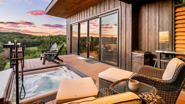 Una terraza de madera con un jacuzzi, mobiliario de mimbre y una puerta de vidrio que da a otra zona de terraza.