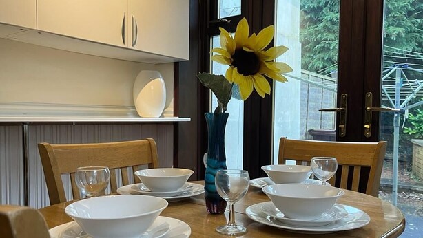 A dining table set with white bowls, plates, and wine glasses, a vase with a sunflower, and wooden chairs.