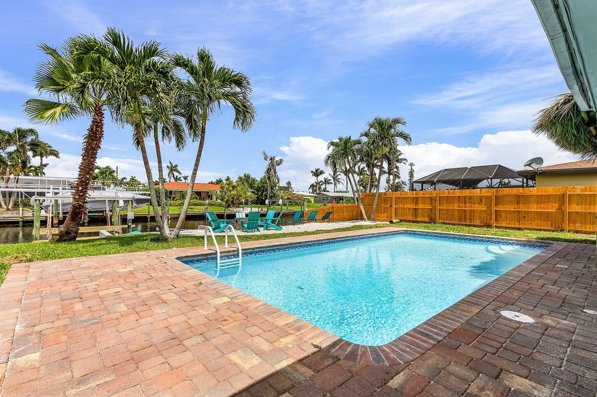 A rectangular swimming pool with a brick border, surrounded by palm trees and a wooden fence.