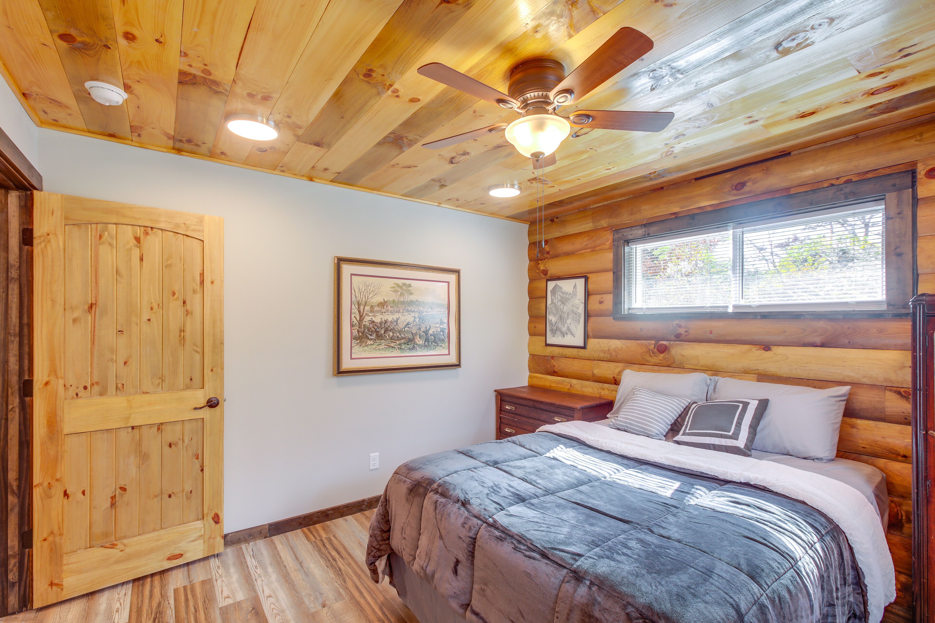 Une chambre dans un chalet en rondins, avec un lit, des murs en bois, un ventilateur de plafond et deux fenêtres.