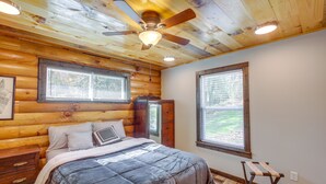 Une chambre avec un plafond en bois, un lit recouvert d’une couette à rayures grises et blanches, une commode en bois et une fenêtre donnant sur un paysage verdoyant.