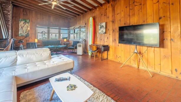 Un salon chaleureux avec un canapé blanc, un téléviseur à écran plat posé sur un pied, des murs en bois et un sol en briques.