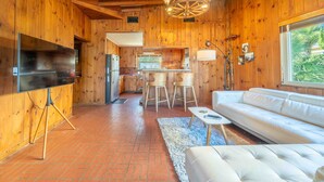 Un intérieur en bois avec un téléviseur posé sur un trépied, un canapé blanc, un coin cuisine et une grande fenêtre.