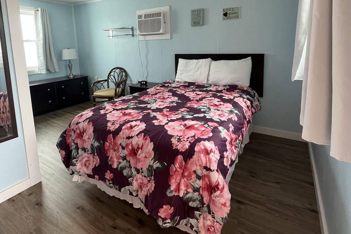 A bedroom with a floral bedspread, a wooden floor, a ceiling fan, and a wall-mounted air conditioner.