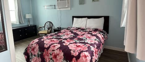 A bedroom with a floral bedspread, a wooden floor, a ceiling fan, and a wall-mounted air conditioner.
