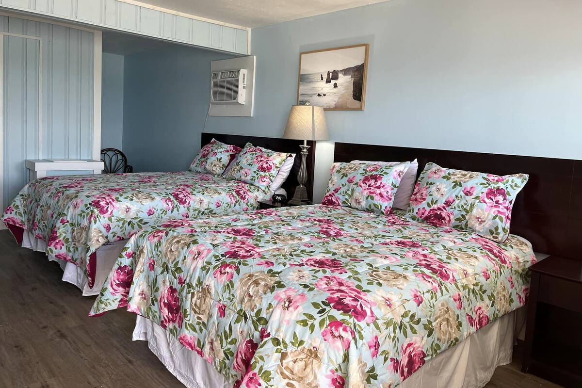 A double room with two beds, each with floral bedding, a bedside table with a lamp, and a framed picture on the wall.