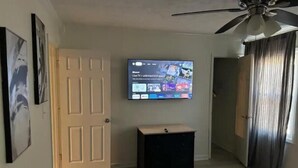 Una habitación con una televisión instalada, una puerta, un mueble negro pequeño y un ventilador de techo.