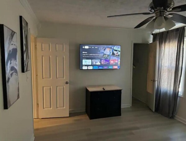 A room with a mounted TV displaying a live TV interface, a door, a small black cabinet, and a ceiling fan.