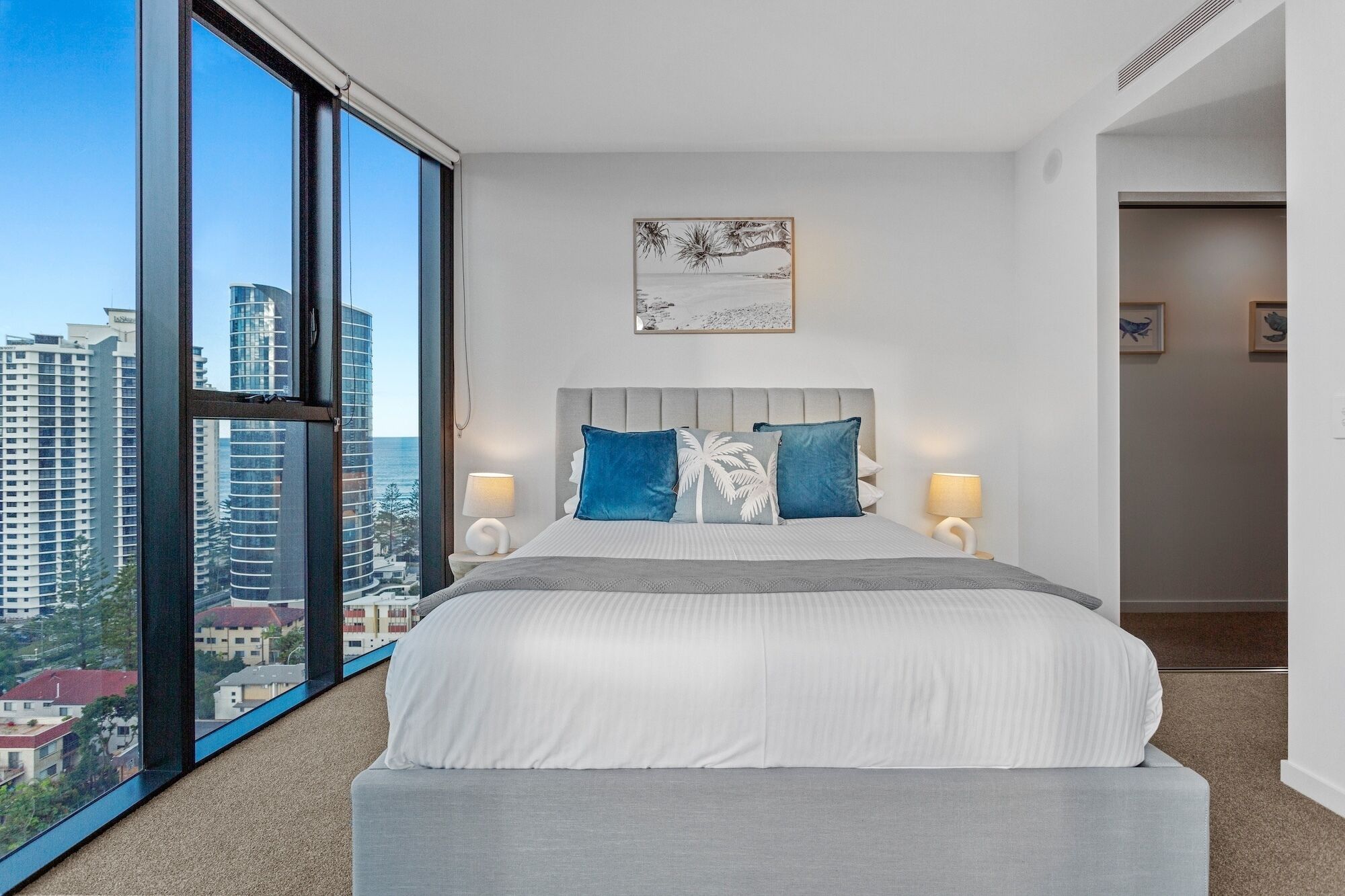 Una habitación de hotel moderna con un ventanal que ofrece vistas a la ciudad, una cama bien hecha con almohadas azules y blancas, y una obra enmarcada sobre la cama.