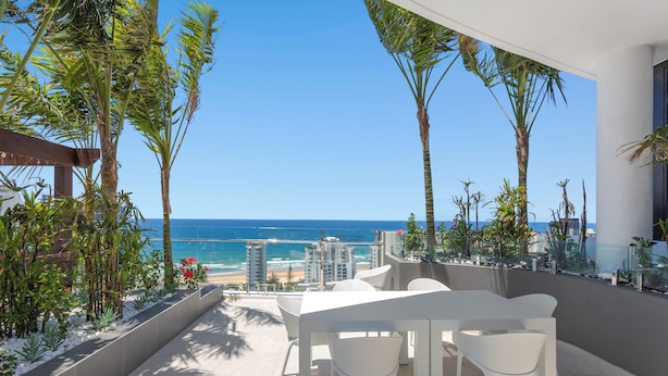 Eine Terrasse mit Blick auf das Meer, weiße Möbel und tropische Pflanzen.