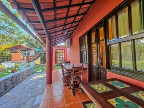 A covered outdoor seating area with a table and chairs, a stone wall, and a view of a garden. - Casa em Geribá (Manguinhos Armação dos Búzios)