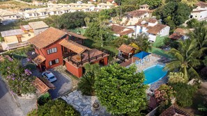 A residential area with a swimming pool, a red building with a balcony, and surrounding greenery. - Casa em Geribá (Manguinhos Armação dos Búzios)