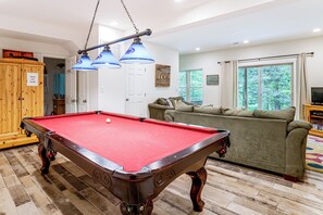 Una mesa de billar con superficie de fieltro rojo, un mueble de madera con un letrero y un sofá verde en una habitación con paredes blancas y un ventanal grande.