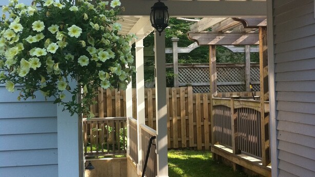 A covered porch with a wooden deck, a hanging flower basket, and a wooden fence in the background. - North Hill Section Home; walk to UVMMC & UVM, Chabad, downtown, lake. (Burlington)