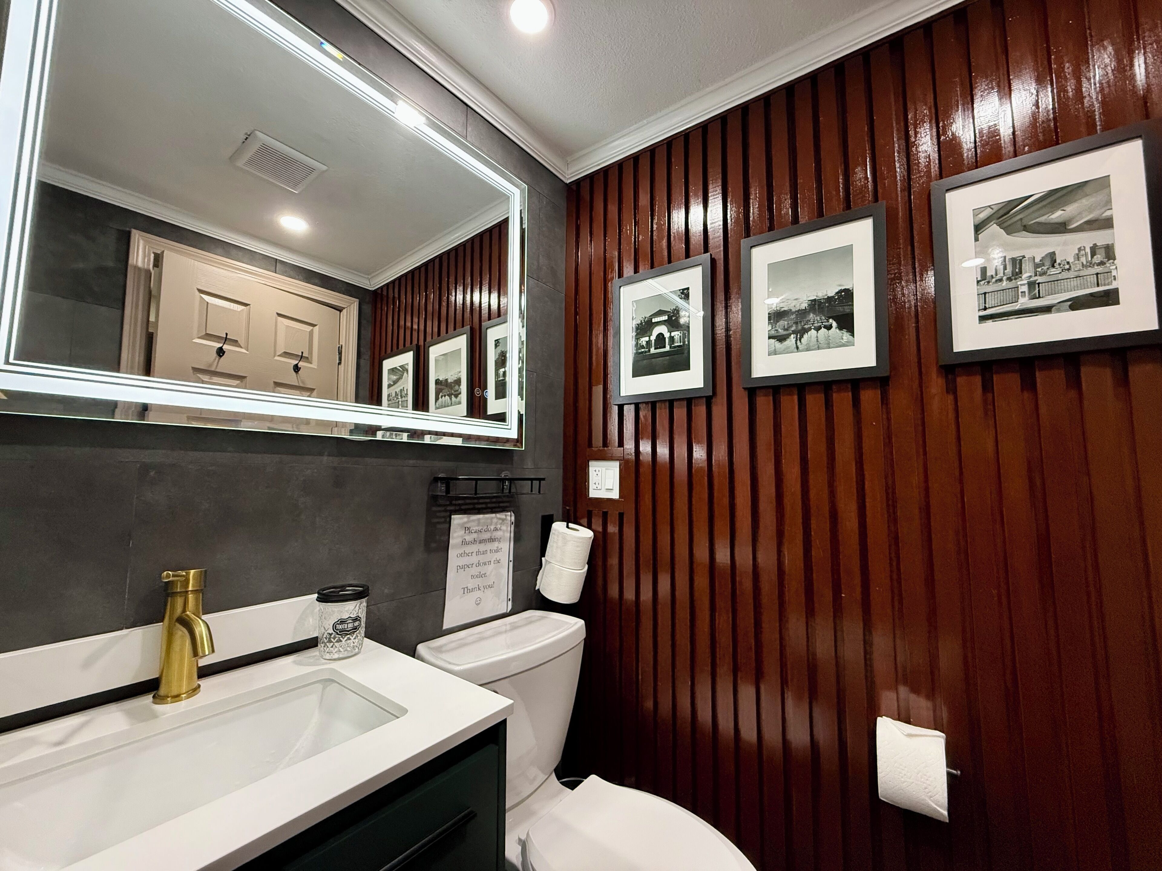 Une salle de bain avec un lavabo blanc, des toilettes et un miroir au-dessus du lavabo. Trois tableaux encadrés sont accrochés au mur.