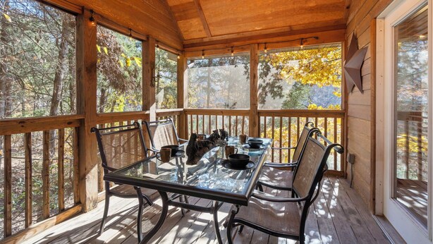 A wooden deck with a glass table and chairs, surrounded by large windows with a forest view. - Little Creek Cabin (Branson West)