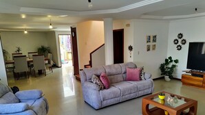 Amplia sala de estar con un sofá gris, una mesa de centro y un televisor. Se ve una escalera que lleva a otra habitación, y un comedor con sillas y una mesa.