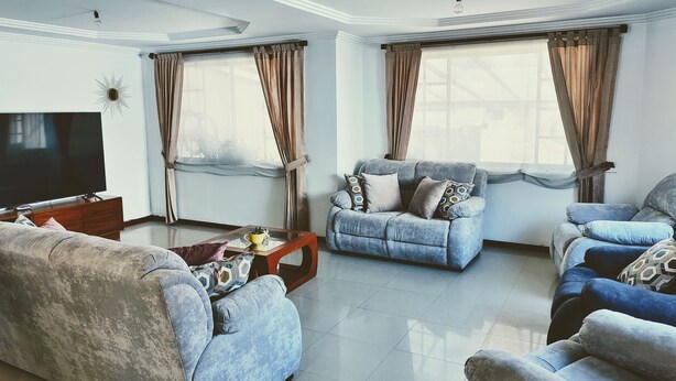 Una sala de estar con televisor de pantalla plana, dos sofás, una mesa de centro y ventanas grandes con cortinas.