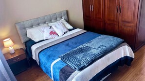 Una cama bien hecha con una colcha azul y blanca, un cabecero gris y un armario de madera al lado.