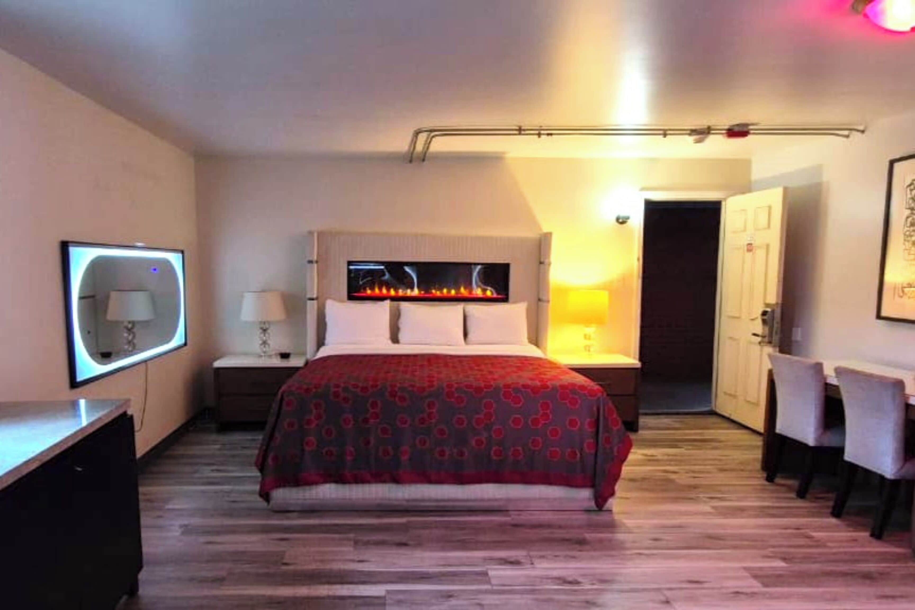 A hotel room with a bed, bedside tables, a TV, a fireplace, and a dining area.