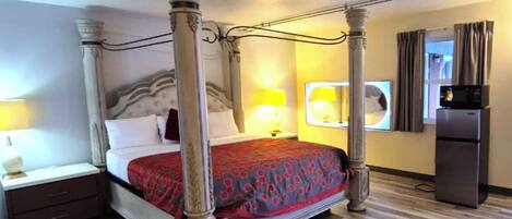 A four-poster bed with a red patterned bedspread, a nightstand with a lamp, a refrigerator, and a window with curtains.