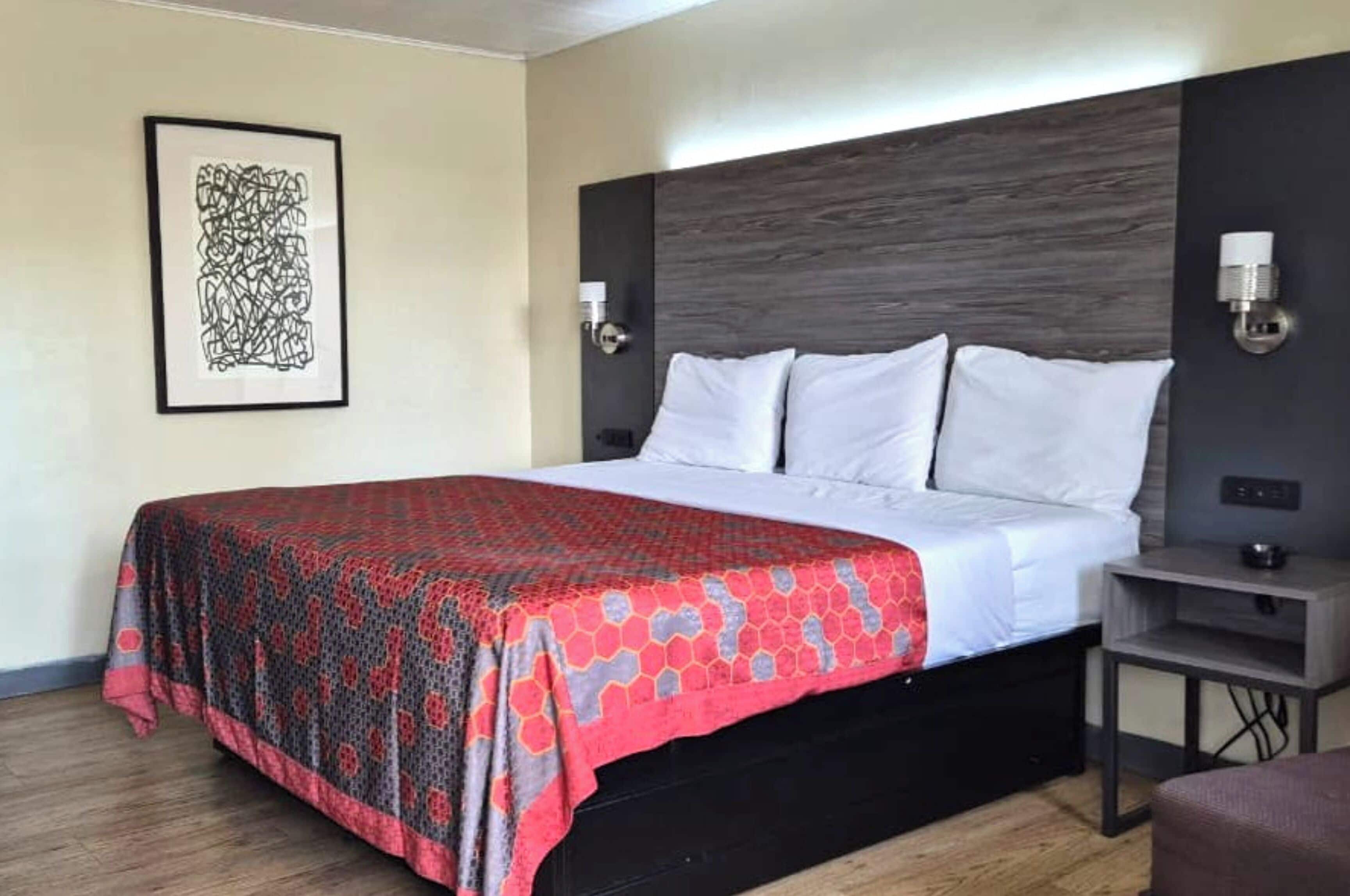 A hotel room with a bed, a headboard, bedside lamps, a framed artwork on the wall, and a small table with a lamp.