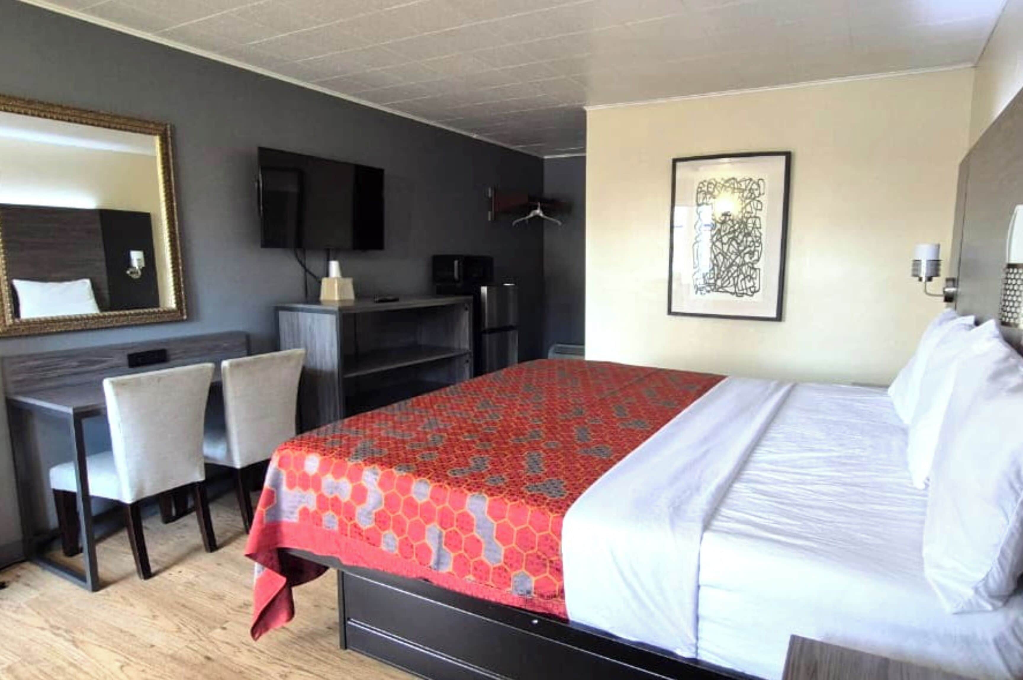 A hotel room with a bed, a desk, two chairs, a television, and a framed artwork on the wall.