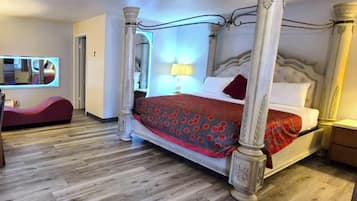 A four-poster bed with a red and brown patterned bedspread, a red pillow, and a white pillow. There is a nightstand with a lamp on it. The room has wooden flooring and a mirror on the wall.