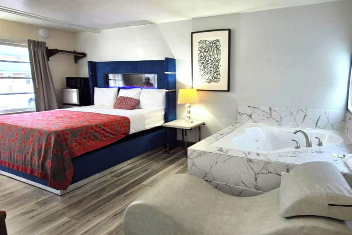 A hotel room with a bed, a large window with curtains, a desk, a lamp, and a bathtub.