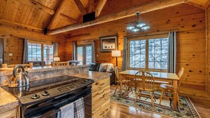 Interior de una cabaña de madera con cocina, comedor y amplios ventanales.