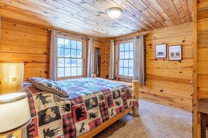 Ein Holzblockhaus-Zimmer mit einem Bett, zwei Fenstern, einer Lampe und gerahmten Bildern an der Wand.