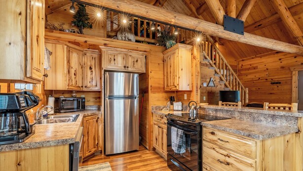 Interior de una cabaña de madera con cocina, armarios de madera, refrigerador de acero inoxidable, microondas, estufa y escalera de madera.